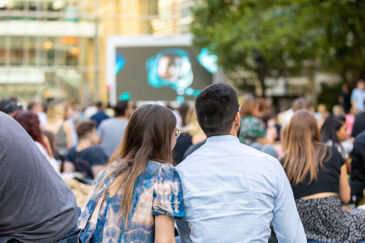 People sitting on the ground watching a large screen, with trees in the background