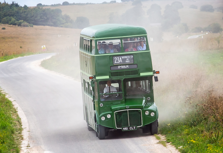 A green double decker bus travelling down an otherwise deserted road with grass and bushes on both sides, kicking up dust as it goes