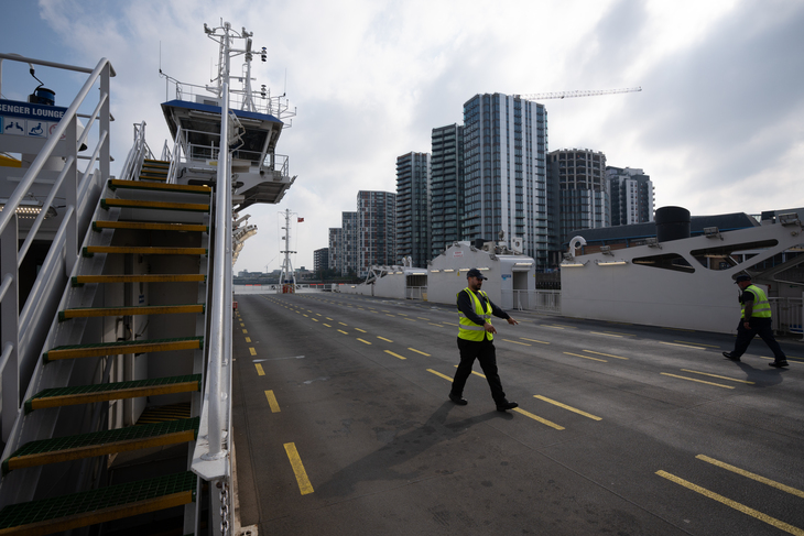 A wide-angle shot from the deck of a ferry looking toward a city skyline of modern high-rise buildings. Two crew members in high-visibility vests walk across the deck, which is marked with yellow lines. A staircase and the ship's bridge are visible on the left.