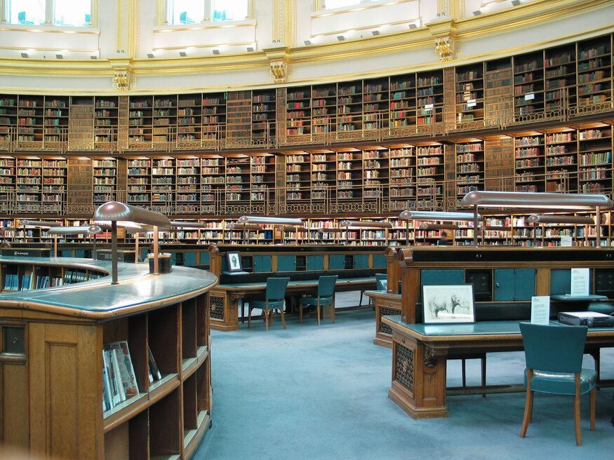 The circular interior of a grand library featuring multi-level, floor-to-ceiling wooden bookshelves, curved study desks with green chairs, and classic desk lamps.