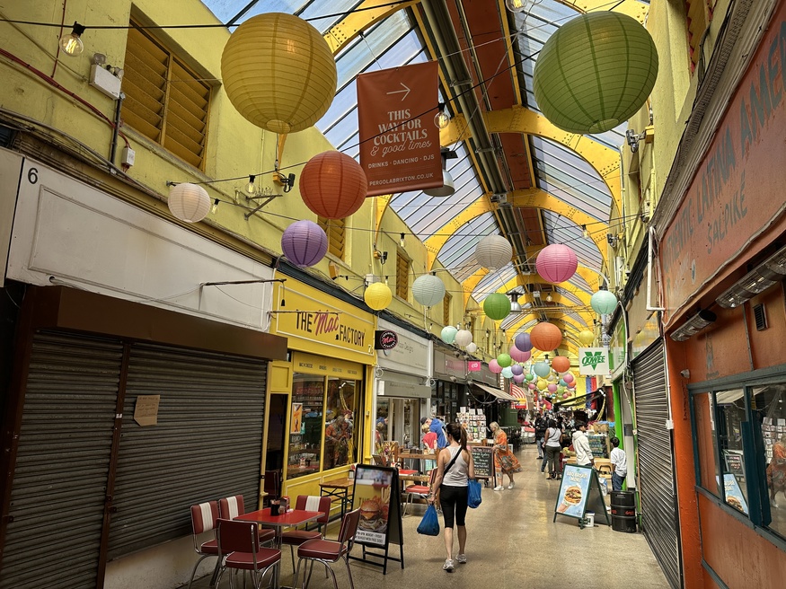 A long, narrow indoor market hallway with a high glass ceiling and yellow walls. Colorful paper lanterns in shades of yellow, green, orange, purple, and pink hang from the ceiling. People walk through the market past storefronts, including a yellow-fronted restaurant called "The Mac Factory" with red chairs and tables outside. A sign overhead points the way to cocktails and dancing.
