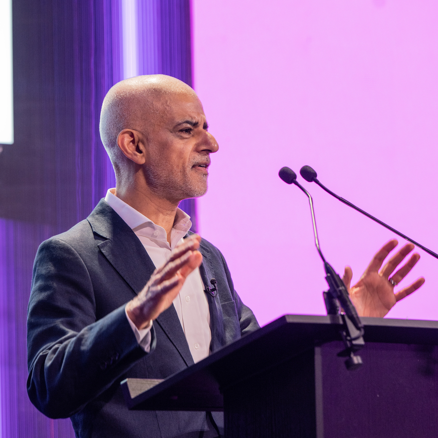 A bald man in a dark blazer and white shirt speaks at a podium with two microphones against a purple background.