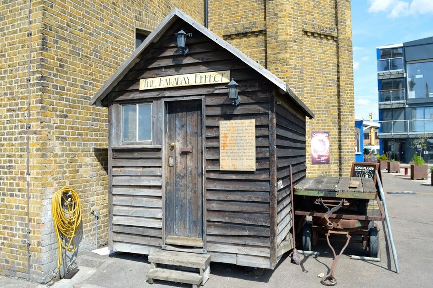 A small, weathered wooden shed labeled "The Faraday Effect" stands against a large brick building, with a wooden cart parked beside it.