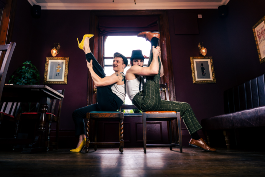 Things to do in London this weekend: two people sitting back to back on chairs, each raising a leg high into the air, creating a symmetrical pose