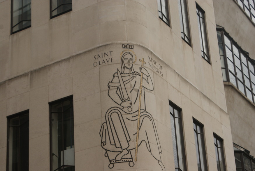 A minimalist mosaic of Saint Olave, King of Norway, on the curved corner of a stone building. The figure is depicted in black and gold outlines, holding a sword and a long cross, with the text "SAINT OLAVE" and "KING OF NORWAY" flanking his head.