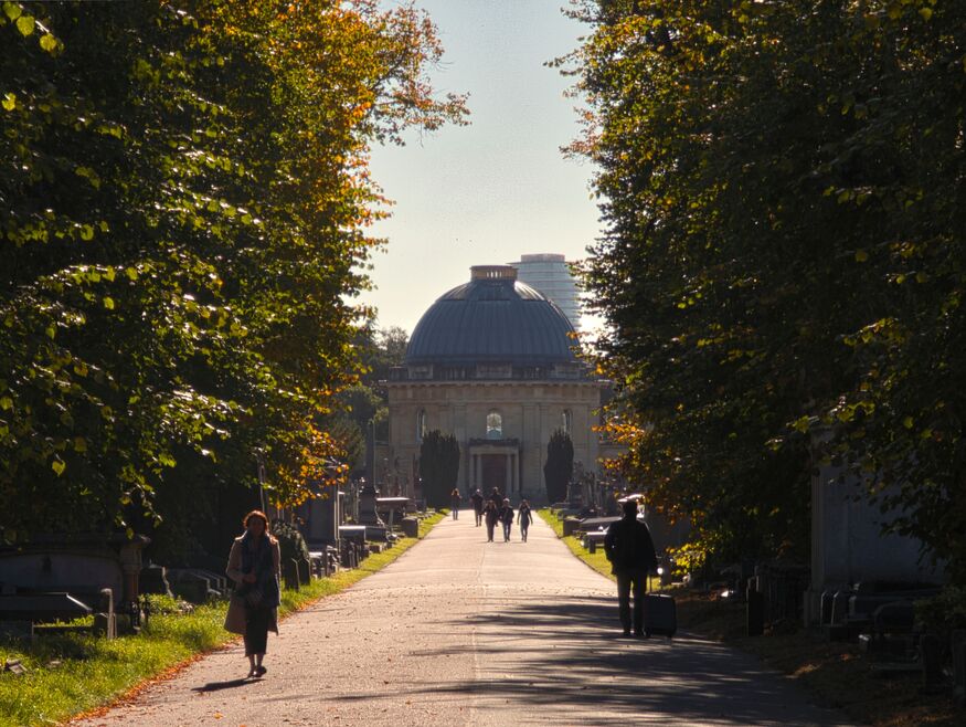 A long, sunlit path through a cemetery lined with tall trees and gravestones leads toward a large domed chapel in the distance, with several people walking along the walkway.