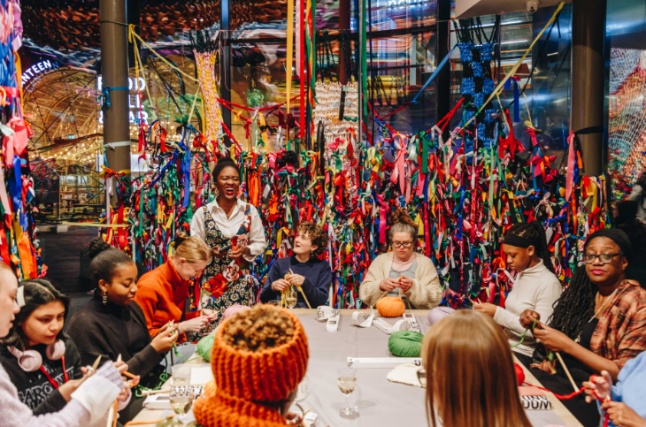 August 2026 in London: a group of people of all ages sitting around a table learning finger knitting