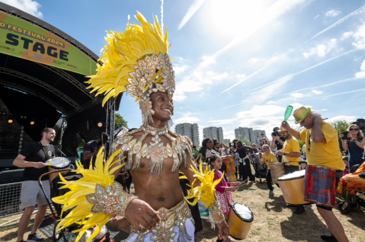 August 2026 in London: a person dressed in a yellow and silver Carnival costume, with drummers in the background, in front of a festival stage