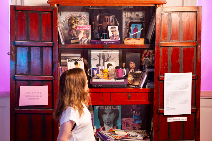 A woman standing in front of a wooden cupboard which is a shrine dedicated to Prince.
