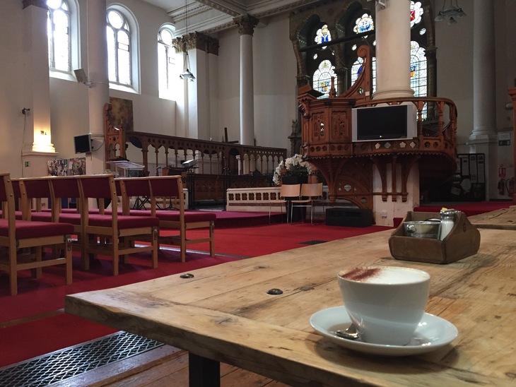 A cup of cappuccino sits on a rustic wooden table in the foreground, with the interior of a church converted into a cafe visible in the background, featuring red carpet, rows of chairs, and an ornate wooden pulpit.