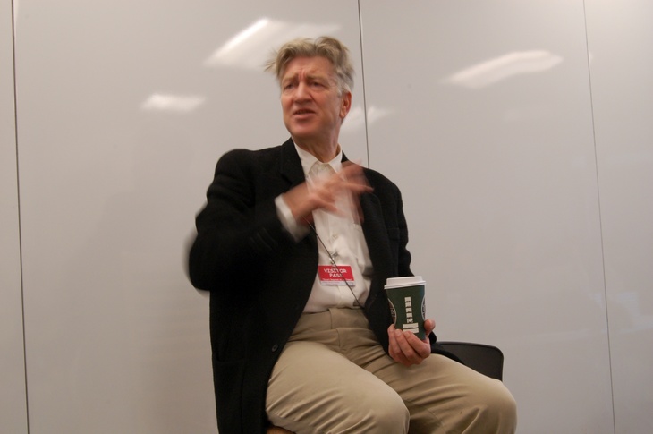 A medium shot of filmmaker David Lynch sitting in a chair against a plain white wall, gesturing with his hand while holding a coffee cup and wearing a visitor pass.