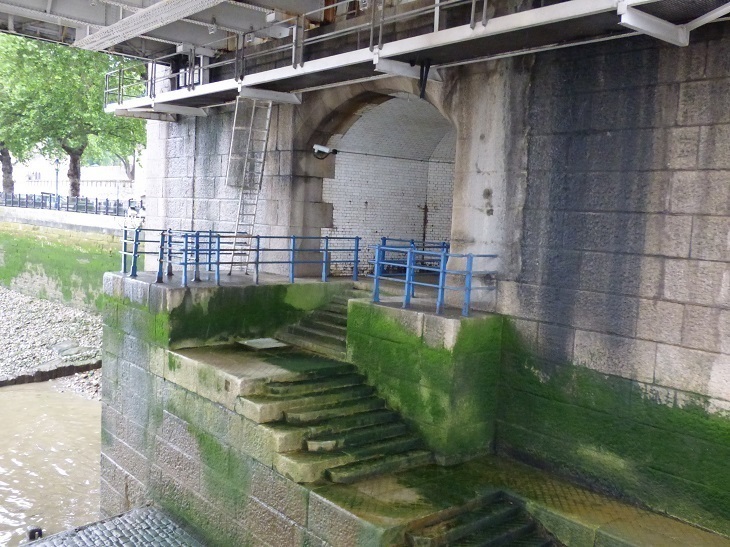 A photo of a set of stairs leading up from the river to a gap in the underside of Tower Bridge