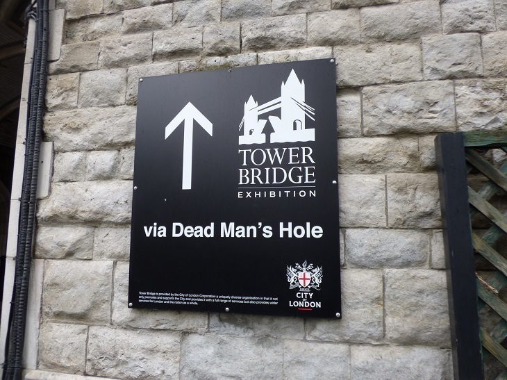 A sign mounted on a wall directing people to Tower Bridge Exhibition via Dead Man's Hole
