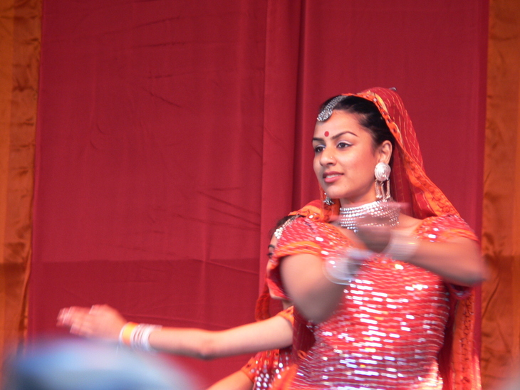 A woman in traditional Indian attire, including an orange sequined dress, jewelry, and a bindi, performs a dance in front of a red curtain.