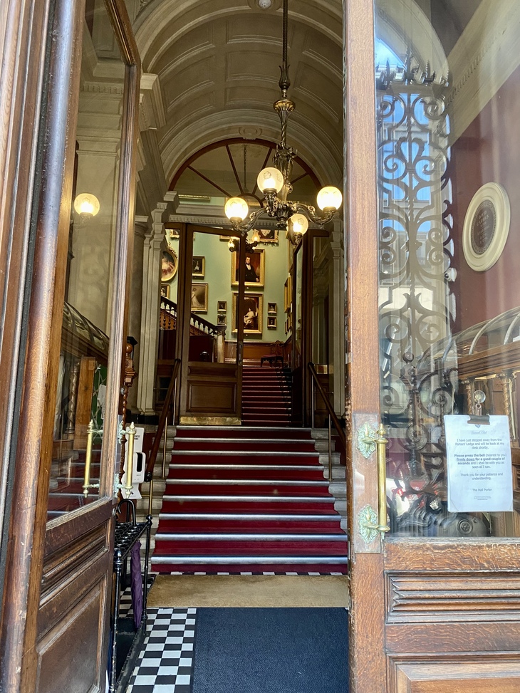 A view through open, ornate wooden doors into a grand, historic interior featuring a red-carpeted staircase, a vaulted ceiling with a brass chandelier, and walls lined with framed portraits.