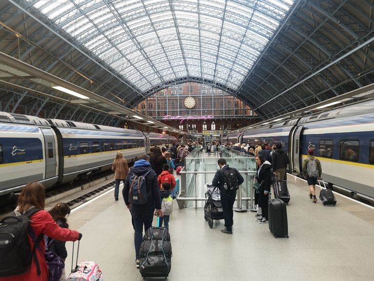 A wide-angle shot of the bustling St Pancras International train station in London, showing passengers with luggage walking along a platform between two Eurostar trains under a large arched glass and steel roof.