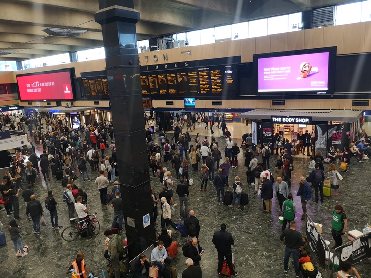 High-angle view of a crowded train station concourse with people standing and walking, large digital departure boards, and retail shops like The Body Shop.
