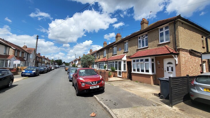 A row of semi-detached houses along a residential street under a blue sky with white clouds, with several cars parked on the road and in driveways.