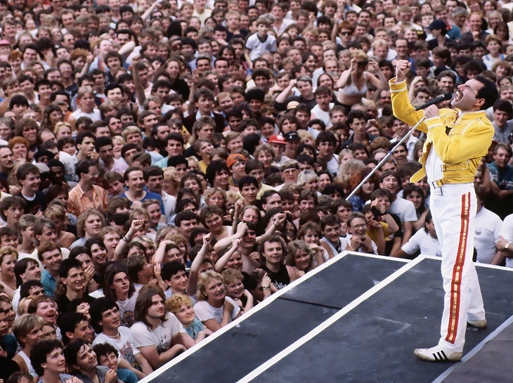 Freddie Mercury, wearing his iconic yellow jacket and white trousers with red stripes, performs on stage before a massive, dense crowd of fans at Wembley Stadium.