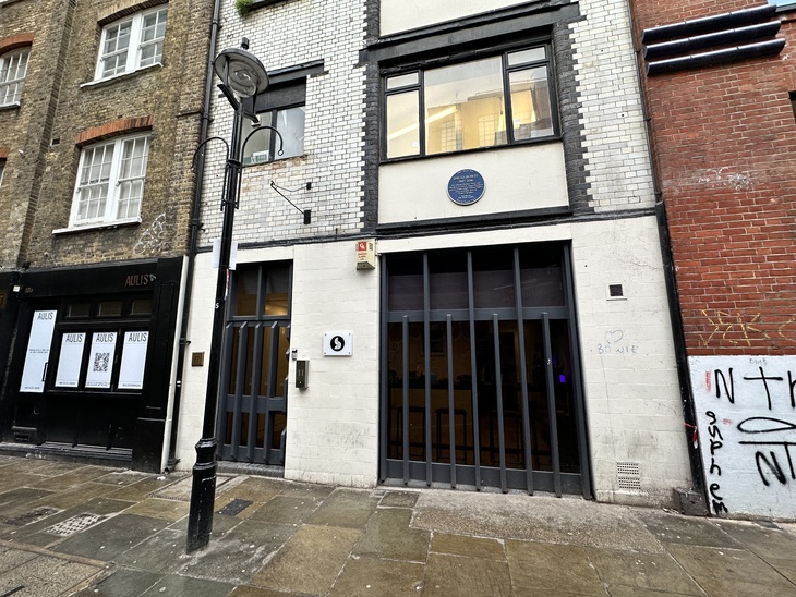 A street-level view of a white brick building in London featuring a blue plaque for David Bowie above a modern storefront with dark grey vertical slats. The building is flanked by a black storefront labeled "AULIS" on the left and a red brick wall with graffiti on the right.