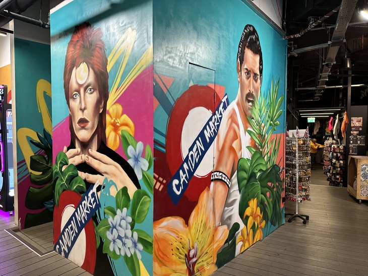 A colorful mural in Camden Market featuring portraits of David Bowie as Ziggy Stardust and Freddie Mercury, surrounded by tropical flowers and London Underground-style signs.