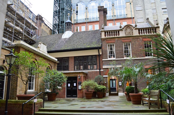 A small, historic brick courtyard in London featuring a mix of architectural styles, with a central gabled building, potted plants, a wooden bench, and modern scaffolding in the background.