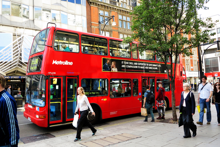 A red double-decker Metroline bus stopped on a busy London street near Bond Street Station, with pedestrians walking on the sidewalk in the foreground.