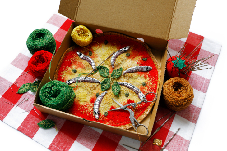 A cardboard pizza box containing a knitted pizza topped with crocheted anchovies. Balls of wool and a tomato-shaped pin cushion sit alongside the box