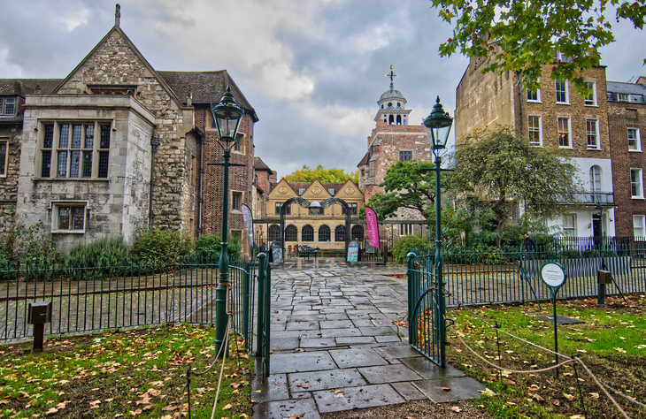 A stone path leads through an open green metal gate toward a courtyard surrounded by historic brick and stone buildings under a cloudy sky. A tall, ornate lamp post stands on each side of the path, and a white cupola is visible in the background.