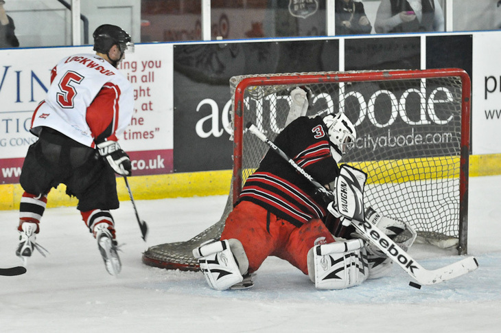 A hockey player in a white and red jersey skates past the goal as the goalie in black and red gear makes a save.
