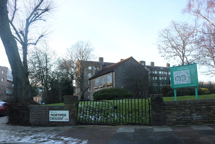 A street sign for Mortimer Crescent NW6 in London, next to a low brick wall and a black metal gate leading to a grassy area with a map of the Mortimer Estate and brick apartment buildings in the background.