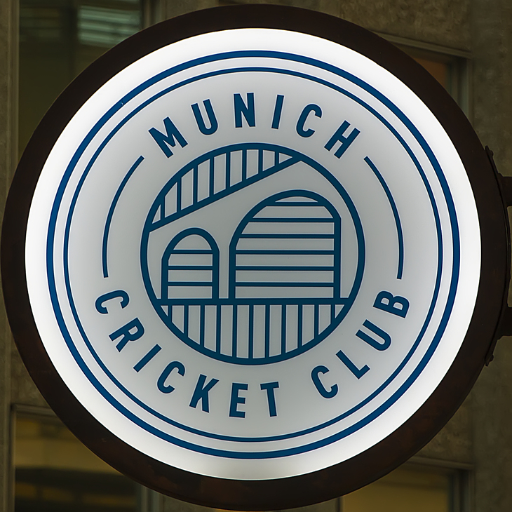A circular, backlit sign for the Munich Cricket Club featuring a blue and white logo with stylized architectural arches and horizontal lines.