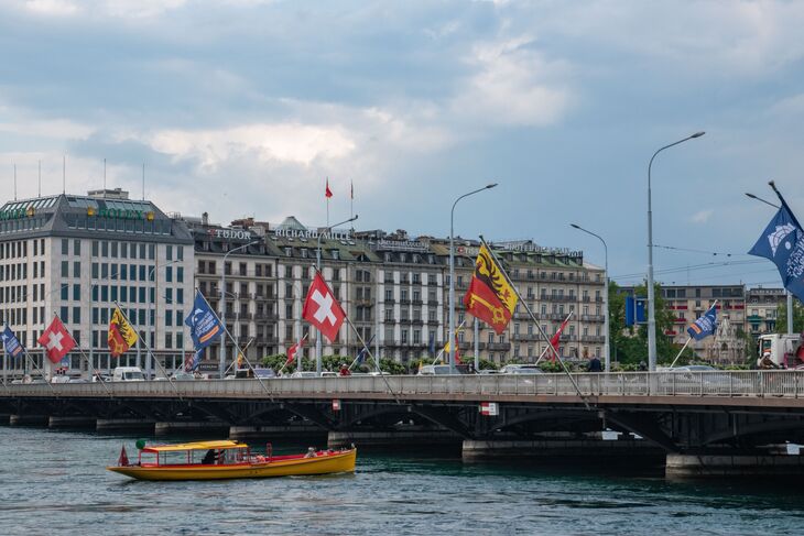 A yellow water taxi cruises on a river in Geneva, Switzerland, in front of the Pont du Mont-Blanc bridge adorned with Swiss and Geneva flags, with luxury watch brand buildings in the background under a cloudy sky.
