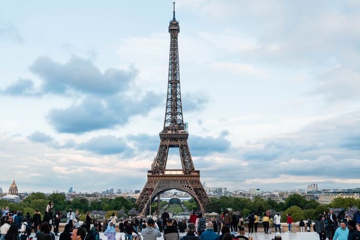 A wide shot of the Eiffel Tower in Paris under a cloudy sky, viewed from a crowded plaza filled with tourists overlooking the city skyline.