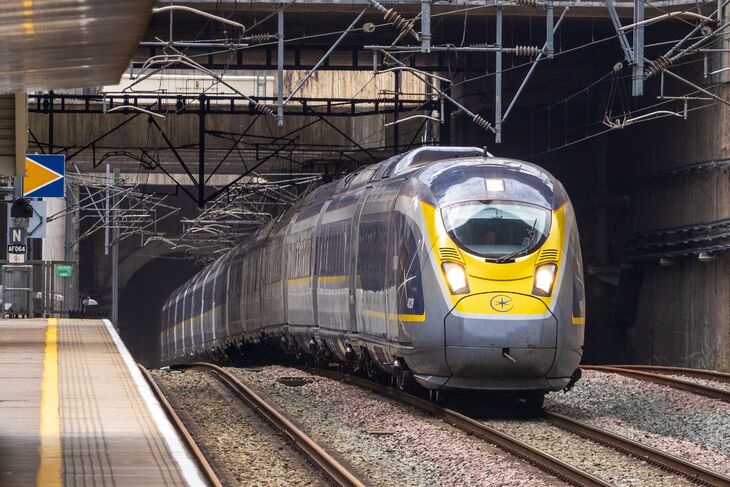 A sleek, modern high-speed Eurostar train with a grey and yellow nose emerges from a dark tunnel onto tracks beside a station platform.