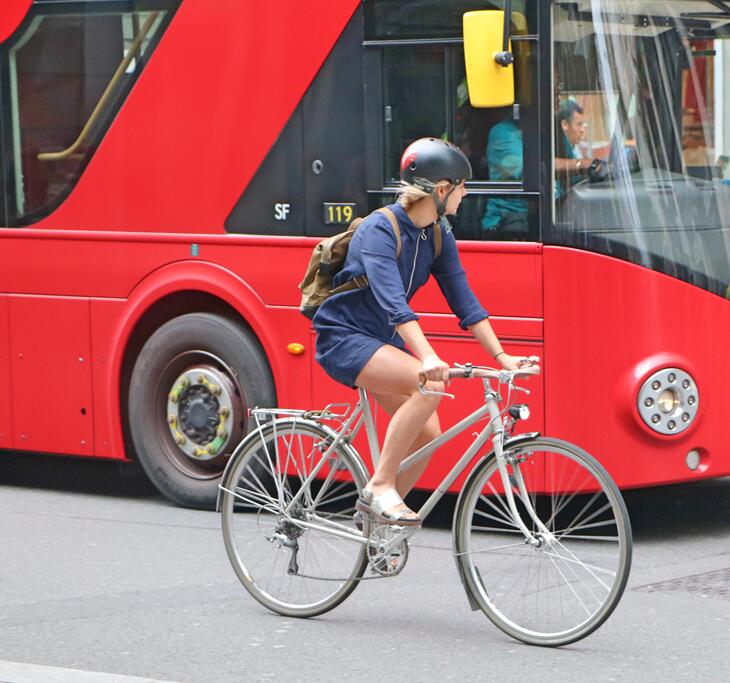 A person in a blue dress and helmet rides a silver bicycle past a large red double-decker bus on a city street.