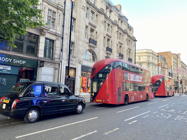 A black London taxi and two red double-decker buses drive down a city street lined with historic stone buildings and shops like The Body Shop and Clarks.