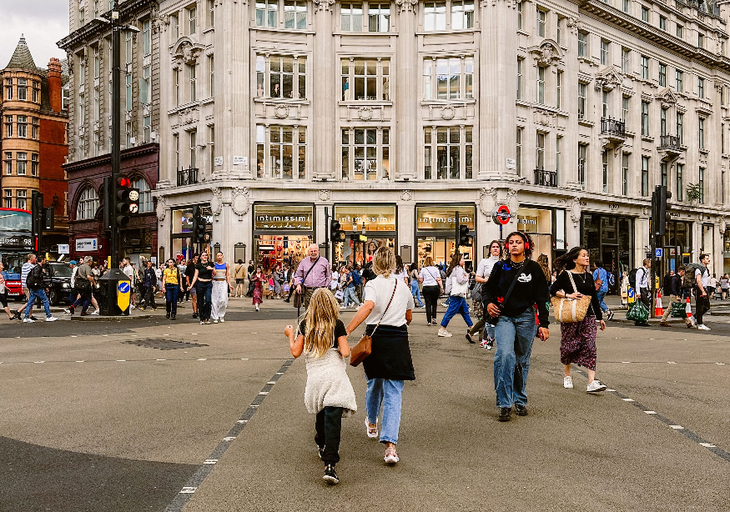 A wide-angle shot of a busy pedestrian crossing at Oxford Circus in London, featuring a large, ornate stone building with storefronts like Intimissimi at its base. Numerous people are walking across the street under a cloudy sky, with a young girl and woman in the foreground walking away from the camera.