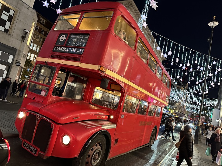A vintage red double-decker bus, labeled "Marble Arch," drives down a London street at night under festive star-shaped lights.