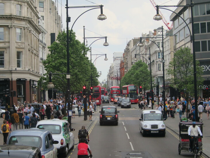 A busy London street scene on Oxford Street, filled with black cabs, red double-decker buses, and crowds of pedestrians on the sidewalks. Street lamps arch over the road, and a rickshaw is visible in the foreground.