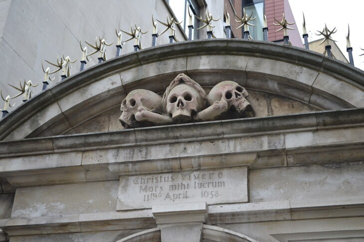 A stone archway featuring three carved skulls and crossbones above a Latin inscription that reads "Christus vivere Mors mihi lucrum 11mo April 1658." The arch is topped with a row of sharp, golden-tipped metal spikes.
