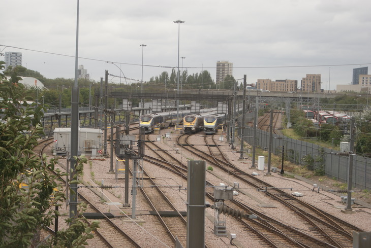 A high-angle view of a railway depot with multiple tracks converging in the foreground. Three silver and yellow Eurostar trains are parked side-by-side under a bridge in the middle ground, with a cityscape and overcast sky in the background.