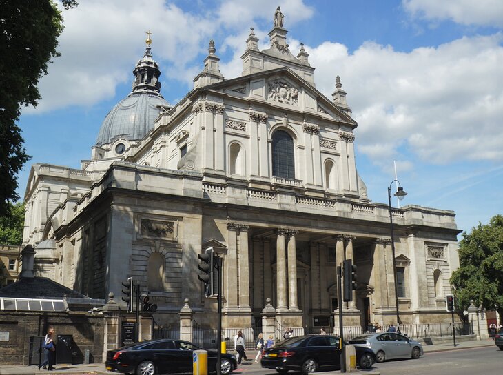 The Brompton Oratory, a large neoclassical Catholic church in London, features a grand stone facade with columns, a prominent dome, and a statue at its peak, set against a blue sky with scattered clouds. In the foreground, cars and pedestrians pass by on a city street.