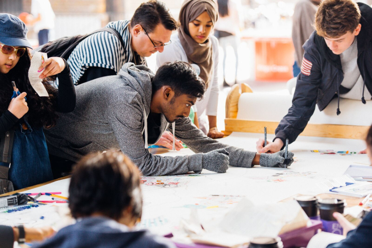Things to do in London in October 2026: people gathered around a large piece of paper on a table, leaning across it and doodling