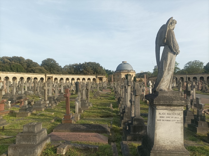 A wide view of Brompton Cemetery in London at dusk, featuring rows of weathered stone headstones and crosses, a large angel statue in the foreground, and a domed chapel in the distance framed by a curved colonnade.