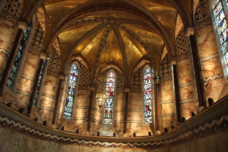 The ornate interior of a chapel featuring a golden mosaic dome, marble walls, and tall stained-glass windows.