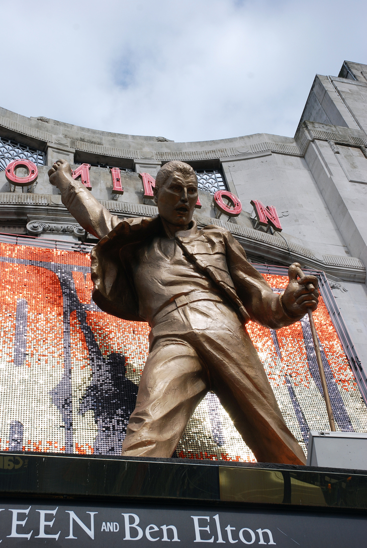 A large gold-colored statue of Freddie Mercury in a triumphant pose stands atop the marquee of the Dominion Theatre in London.