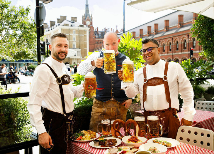 Things to do in London in October 2026: three people standing outside the German Gymnasium wearing traditional Oktoberfest costumes, with steins of beer and pretzels