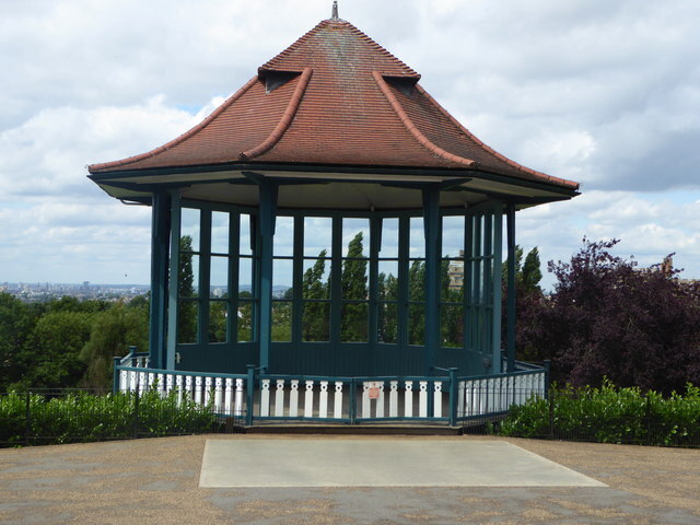 An octagonal blue bandstand with a red tiled roof stands on a hilltop overlooking a city skyline under a cloudy sky.
