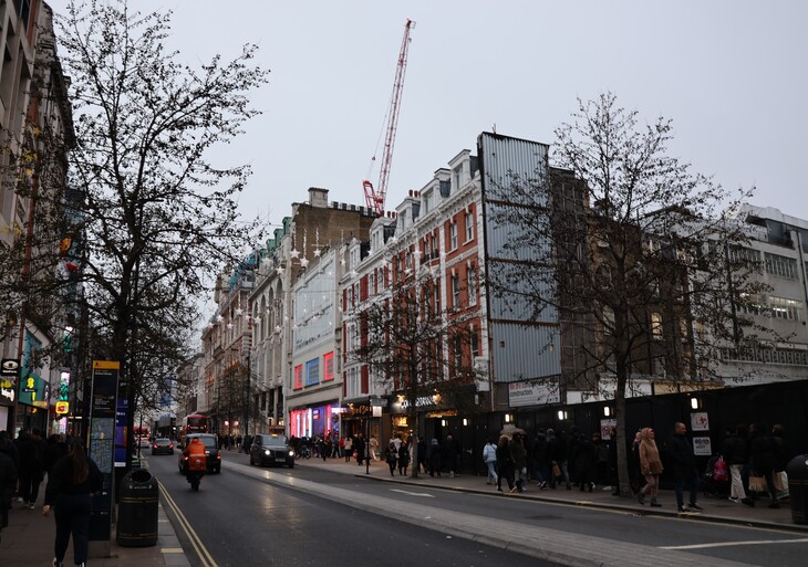 A busy city street at dusk with pedestrians on the sidewalks, cars and a black taxi on the road, and historic buildings decorated with star-shaped lights under a grey sky.